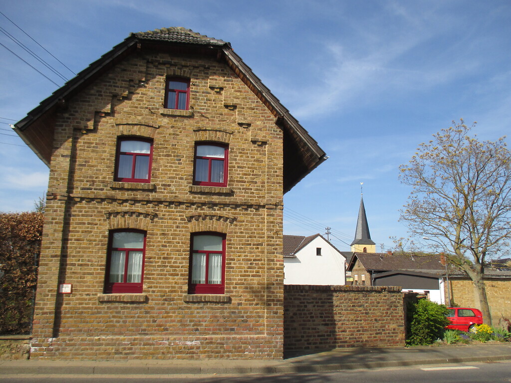 Dieses giebelständige Haus in Muldenau mit Rundbogenfenstern weist eine Backsteinfassade mit Verzierungen auf, im Hintergrund ist der Turm der Pfarrkirche St. Barbara zu sehen. (2015)