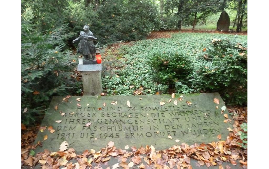 Gedenkstätte für Opfer der NS-Herrschaft im Gremberger Wäldchen (2014). Inschrift auf der Gedenkplatte. Neben der Platte steht die kleine Skulptur, im Hintergrund schließen sich grüne Bodendecker an. Die Fläche wird von Koniferen begrenzt.