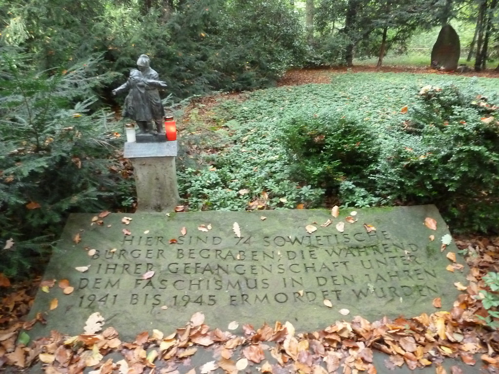 Gedenkstätte für Opfer der NS-Herrschaft im Gremberger Wäldchen (2014). Inschrift auf der Gedenkplatte. Neben der Platte steht die kleine Skulptur, im Hintergrund schließen sich grüne Bodendecker an. Die Fläche wird von Koniferen begrenzt.