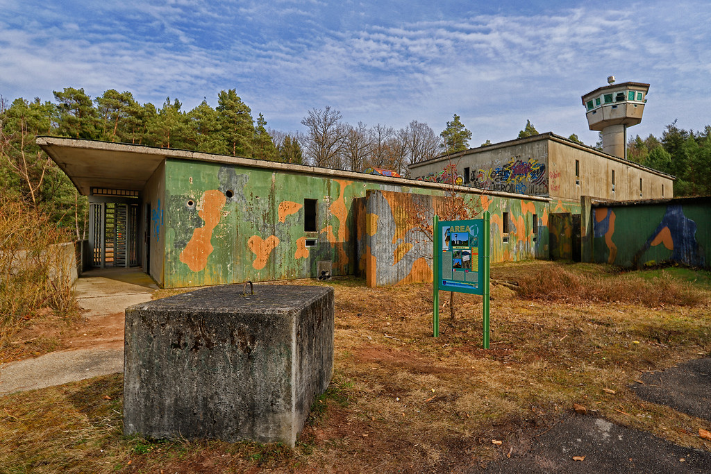 Leerstehende Gebäude des ehemaligen US-Sonderwaffenlagers der Area One in Fischbach bei Dahn. Rechts im Bild steht der ehemalige Überwachungsturm. Im Vordergrund ist eine der heute auf dem Gelände aufgestellten Informationstafeln zu sehen (2018).