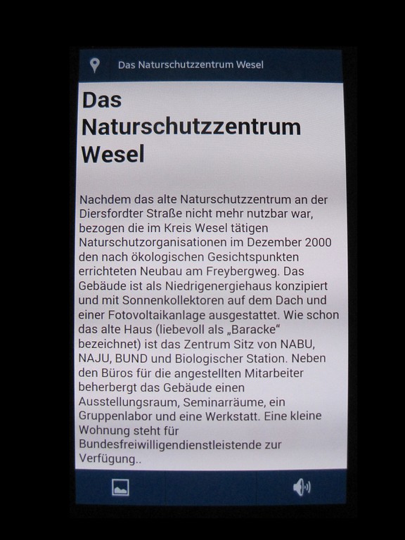 Bildschirm-Foto eines Mobilgerätes von der Anwendungssoftware "App in die Natur!". Es ist den Naturlehrpfad durch das Naturschutzgebiet Weseler Aue und zeigt den Text für den Informationspunkt "Das Naturschutzzentrum Wesel" (2014).