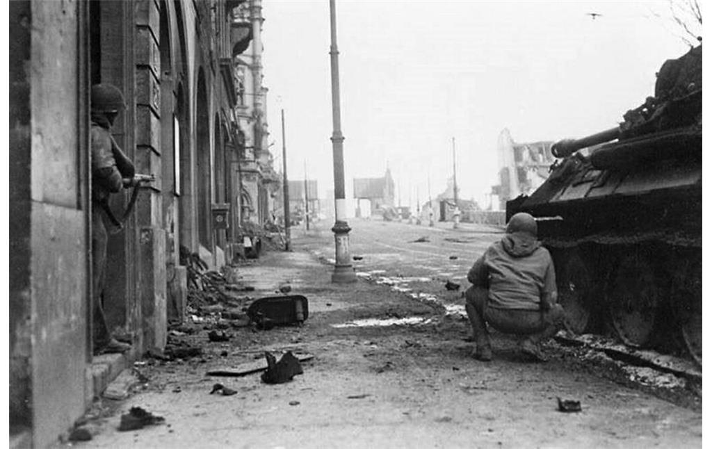 Aufnahme aus den letzten Tagen des Zweiten Weltkriegs in Bonn 1945: mit einem Panzerfahrzeug vorrückende alliierte Soldaten im Bereich der Brückenstraße und der am 8. März 1945 gesprengten Alten Rheinbrücke.