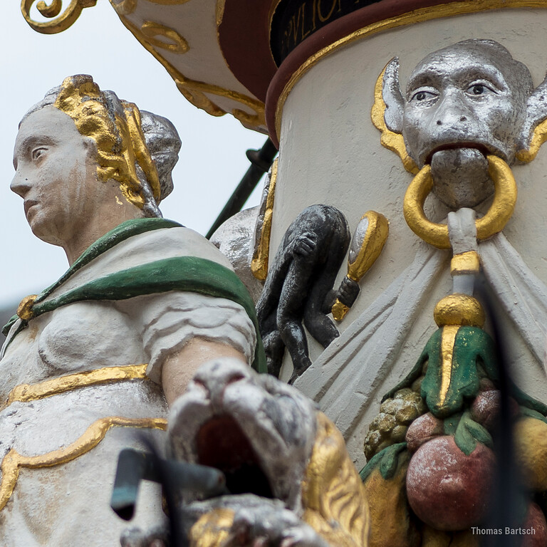 Detail am Petrusbrunnen auf dem Hauptmarkt in Trier (2022): Vier Statuen am Sockel stellen die Kardinaltugenden dar, hier links im Bild die Fortitudo (Tapferkeit und Stärke). Hinter ihrem Rücken am Brunnenstock betrachtet ein Affe sein Hinterteil im Spiegel.