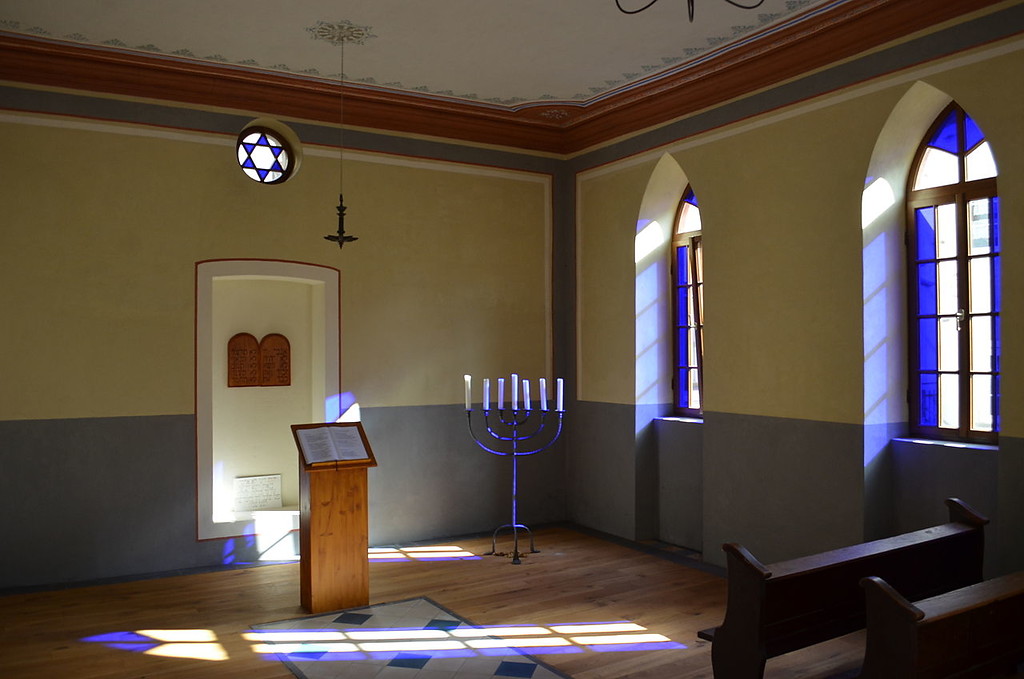 Innenansicht der Synagoge Ediger-Eller mit Lesepult, siebenarmigem Leuchter und buntverglasten gotischen Fensteröffnungen (2015).