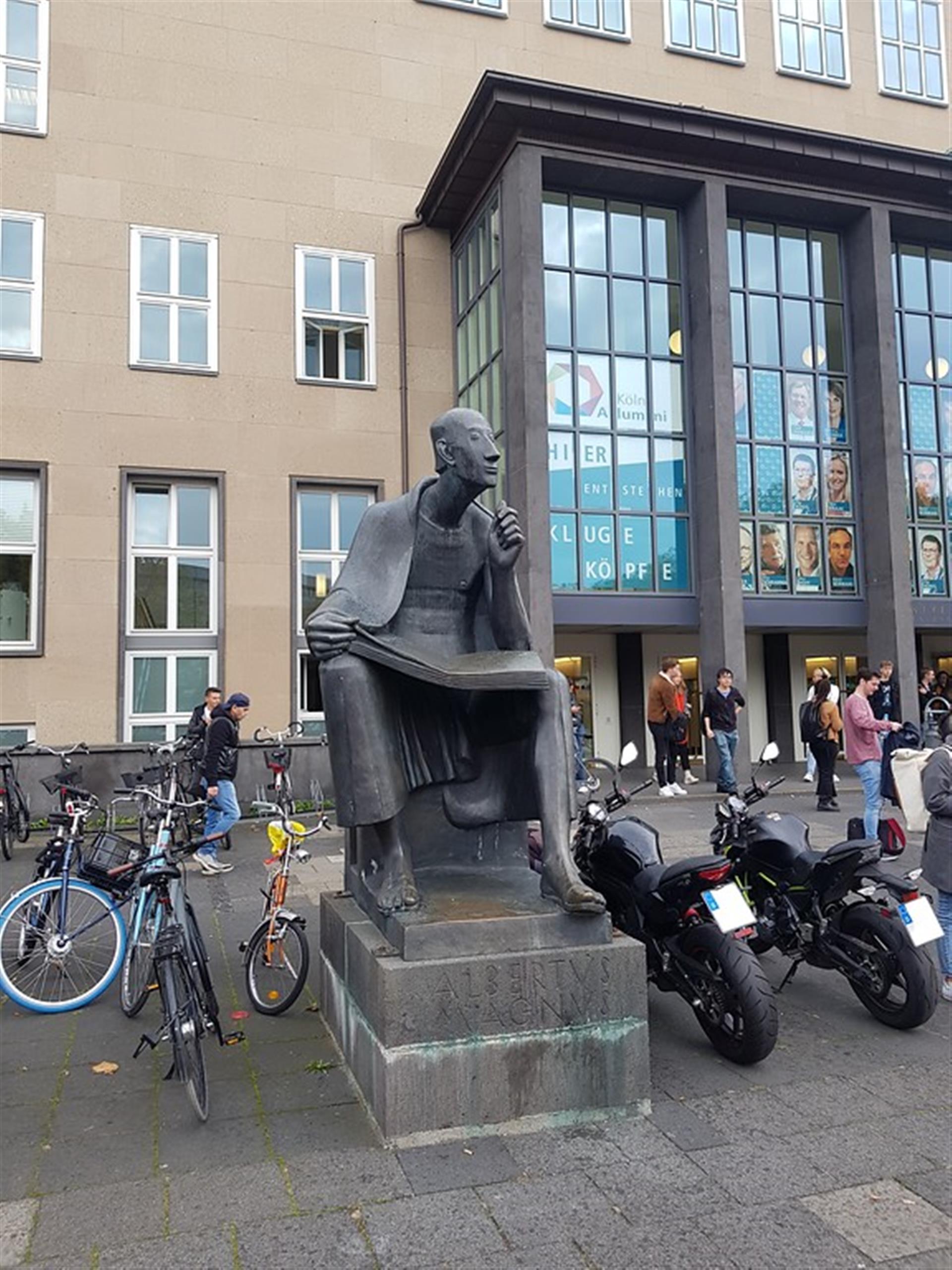In der Bildmitte die Albertus-Magnus-Statue. Im Hintergrund der Eingangsbereich des Hauptgebäudes der Universität zu Köln. Links und rechts von der Statue stehen Fahrräder und Motorräder (2019). &copy; Lennert Herden / Landschaftsverband Rheinland