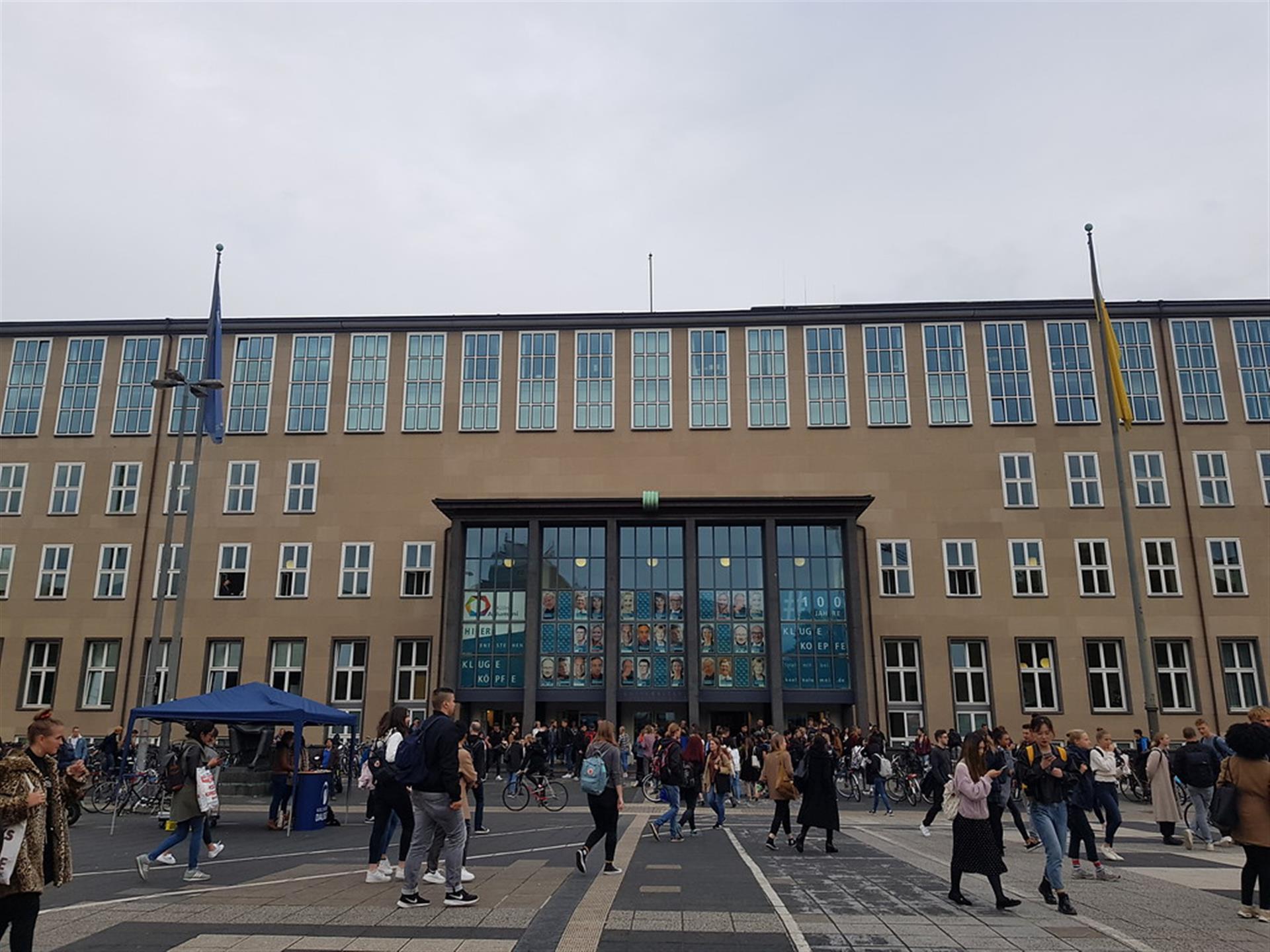Frontalansicht des Hauptgebäudes der Universität zu Köln bei bedecktem Himmel. Der gläserne Eingangsbereich ist in der Mitte zu sehen, jeweils auf der rechten und linken Seite des Einganges befinden sich Fahnenmasten. Es sind viele Menschen vor dem Gebäude auf dem Platz unterwegs (2019). &copy; Lennert Herden