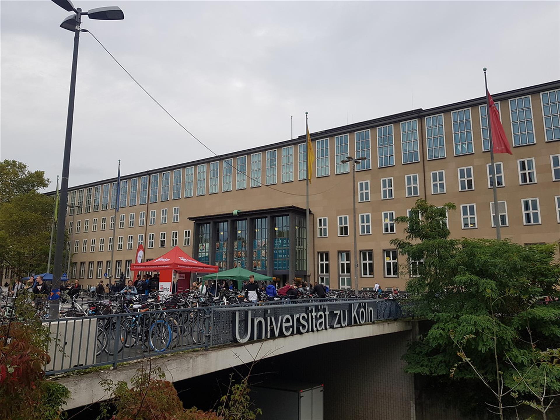 Das Hauptgebäude der Universität zu Köln bei bedecktem Himmel. Es ist der Schriftzug der Universität auf dem Geländer des Albertus-Magnus-Platzes zu erkennen. Es sind am Bildrand Bäume zu erkennen (2019). &copy; Lennert Herden