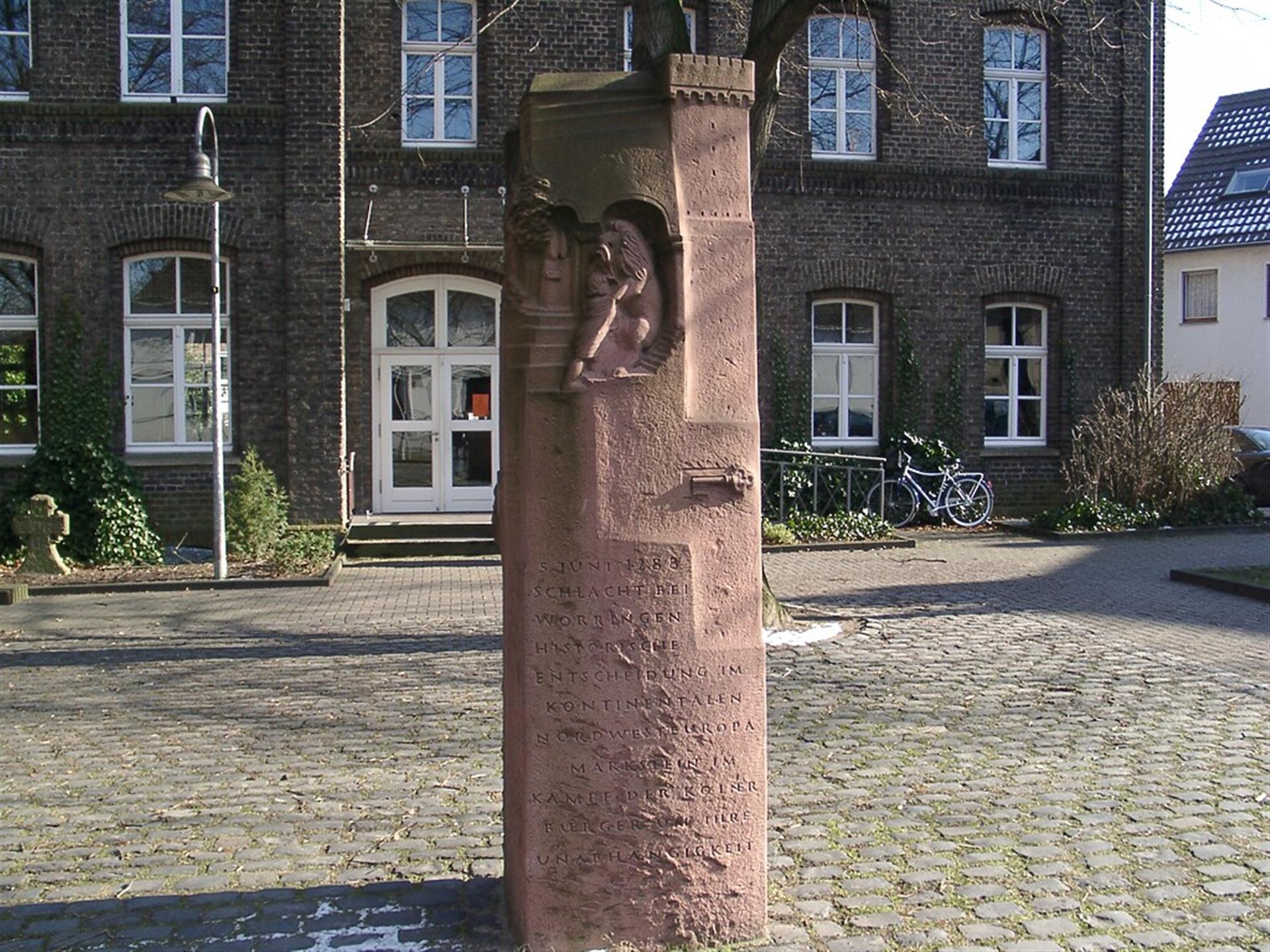 Denkmal zur Erinnerung an die Schlacht von Worringen 1288 in Köln-Worringen (2005). Oben ist ein Mann dargestellt, der mit einem Löwen kämpft. Darunter ist ein Schlüssel zu sehen. Auf dem Denkmal steht geschrieben: "5. JUNI 1288, SCHLACHT BEI WORRINGEN, HISTORISCHE ENTSCHEIDUNG IM KONTINENTALEN NORDWESTEUROPA MARKSTEIN IM KAMPF DER KÖLNER BÜRGER UM IHRE UNABHÄNGIGKEIT" &copy; Klaus Graf / gemeinfrei (via Wikimedia Commons)