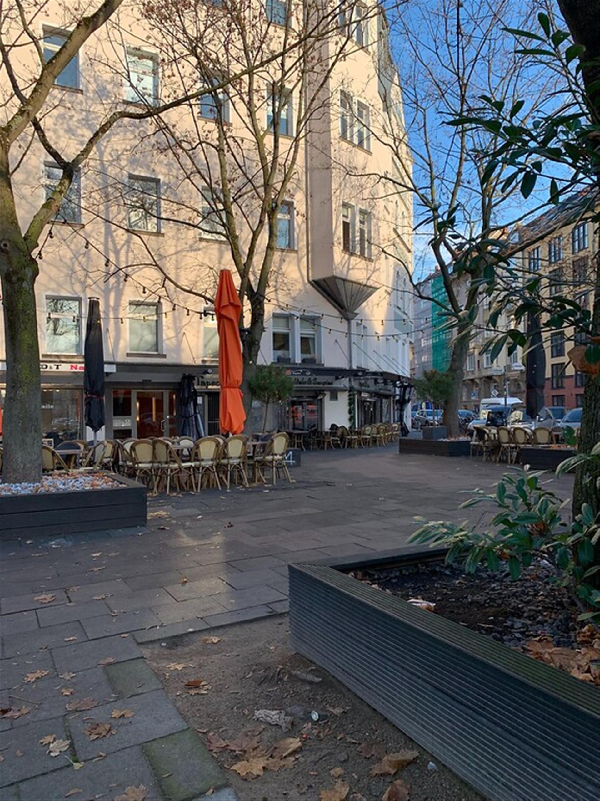 Der Jean-Claude-Letist-Platz in Köln-Altstadt-Süd (2021). Auf dem Platz befinden sich Sitzmöglichkeiten eines Cafés, ebenso wie die Hochbeete einiger Bäume, deren Ränder als Bänke benutzt werden können. &copy; Janina Schwiderski / Landschaftsverband Rheinland / CC-BY-4.0