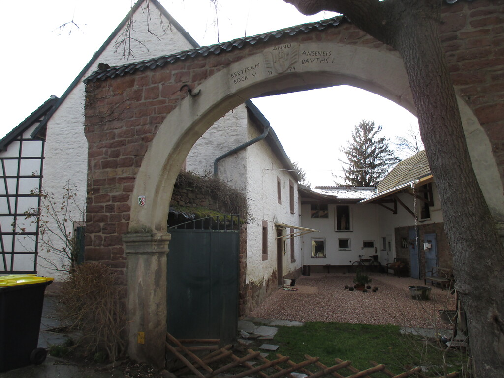 Der Zugang zu dem Hof eines weiß gestrichenen Bruchsteingebäudes führt durch einen Rundbogen aus Sandstein mit Wappen und Inschrift. (2015)