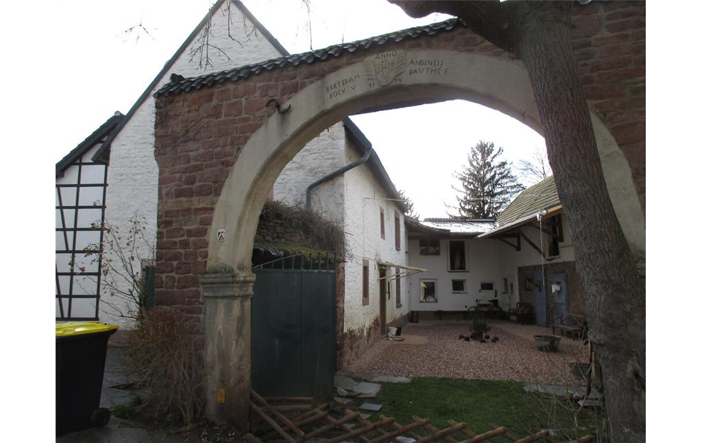 Der Zugang zu dem Hof eines weiß gestrichenen Bruchsteingebäudes führt durch einen Rundbogen aus Sandstein mit Wappen und Inschrift. (2015)