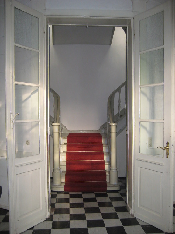 Das historische Treppenhaus des Bahnhofs Belvedere in Müngersdorf (2010). Weiße hohe Holztüren mit Glasfenstern öffnen sich zum Treppenhaus. Die graue, doppelläufige Holztreppe hat ein gedrechseltes Geländer und einen roten Teppich auf den Stufen. Der Boden ist mit schwarzen und weißen Fliesen im Mosaikmuster ausgelegt.