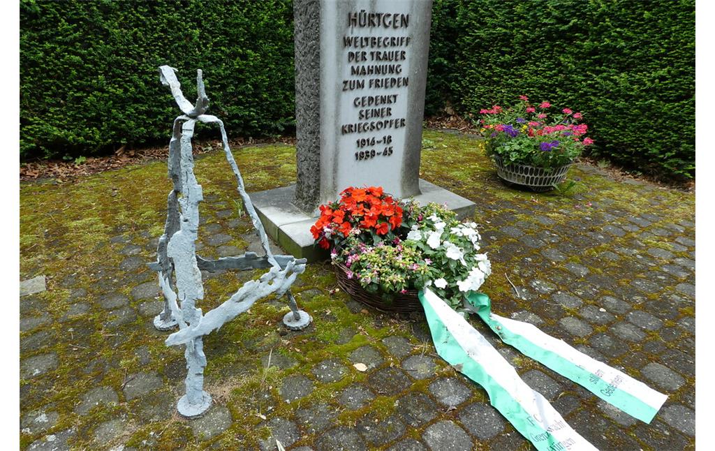 Bild 3: Ein weiß gestrichener Kreuzständer aus Bomben- und Granatsplittern. Dieser steht links neben dem Mahnmal auf dem Gemeindefriedhof Hürtgen (Aufnahme vom 13.07.2019).