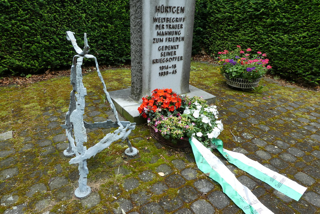 Bild 3: Ein weiß gestrichener Kreuzständer aus Bomben- und Granatsplittern. Dieser steht links neben dem Mahnmal auf dem Gemeindefriedhof Hürtgen (Aufnahme vom 13.07.2019).