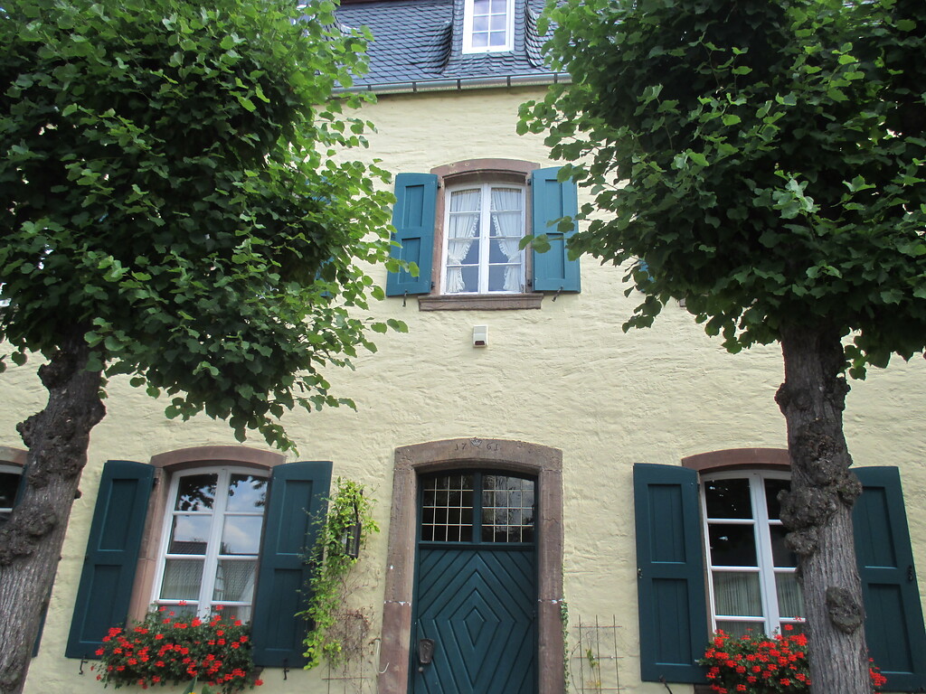 Haus in Embken mit in rotem Sandstein gefassten Rundbogenfenstern und grünen Schlagläden. Die Eingangstür ist ebenfalls aus grünem Holz mit Muster, Rundbogen und Oberlicht, die Einfassung besteht auch aus Sandstein. Davor befindet sich eine Reihe Linden. (2015)