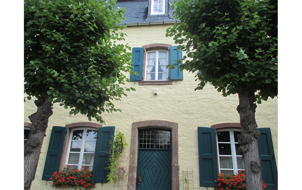 Haus in Embken mit in rotem Sandstein gefassten Rundbogenfenstern und grünen Schlagläden. Die Eingangstür ist ebenfalls aus grünem Holz mit Muster, Rundbogen und Oberlicht, die Einfassung besteht auch aus Sandstein. Davor befindet sich eine Reihe Linden. (2015)