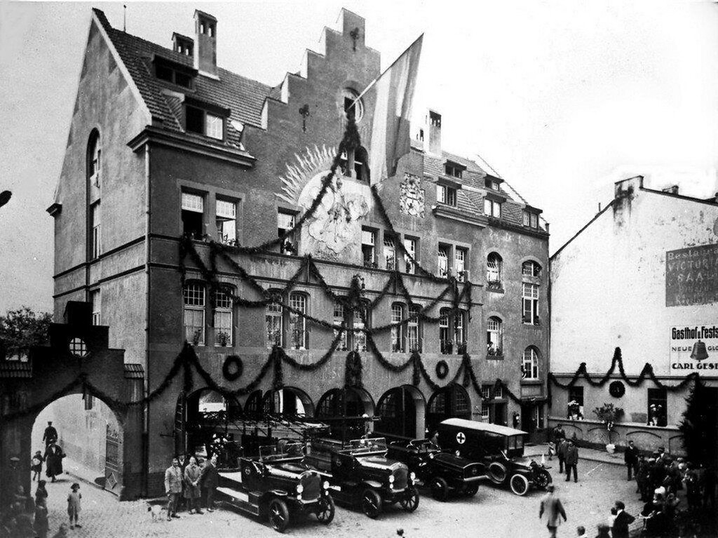 Historische Aufnahme der festlich geschmückten Alten Feuerwache Bonn in der damaligen Karl-Marx-Straße (9. August 1924), davor drei Feuerwehrfahrzeuge und ein Krankenwagen.