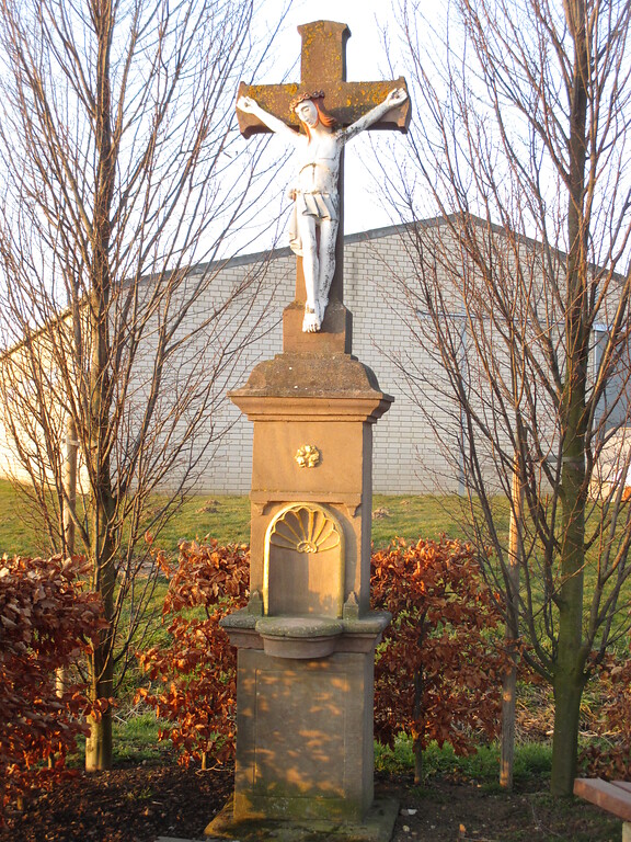 Wegekreuz an einer Weggabelung bei Füssenich. Das Rotsandsteinkreuz mit Korpus aus Stein steht auf einem untergliederten Sockel mit Nische und goldenen Verzierungen. (2015)