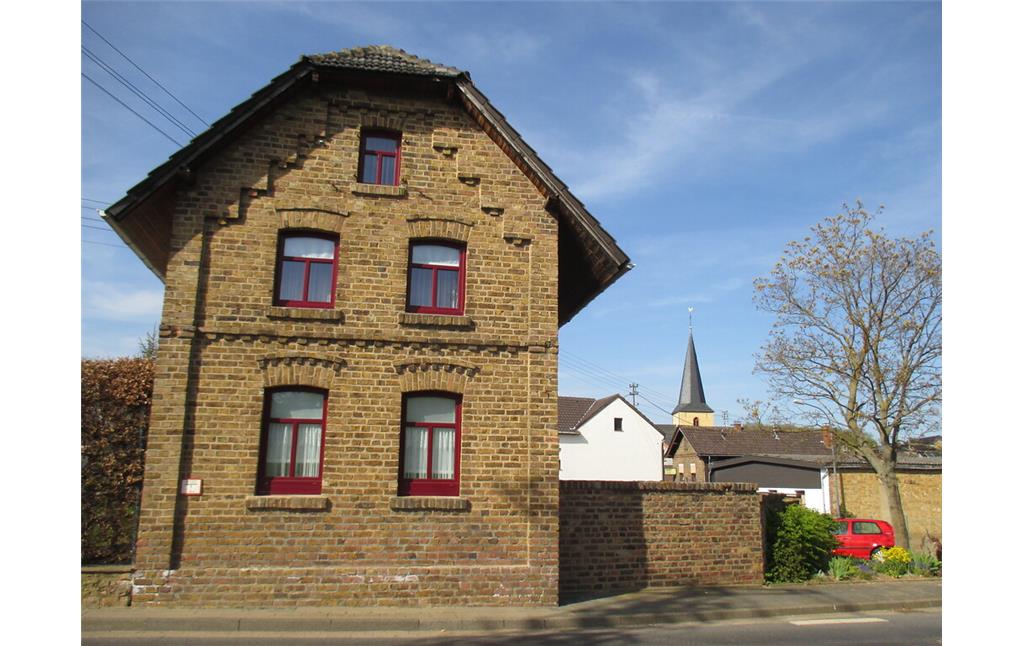 Dieses giebelständige Haus in Muldenau mit Rundbogenfenstern weist eine Backsteinfassade mit Verzierungen auf, im Hintergrund ist der Turm der Pfarrkirche St. Barbara zu sehen. (2015)