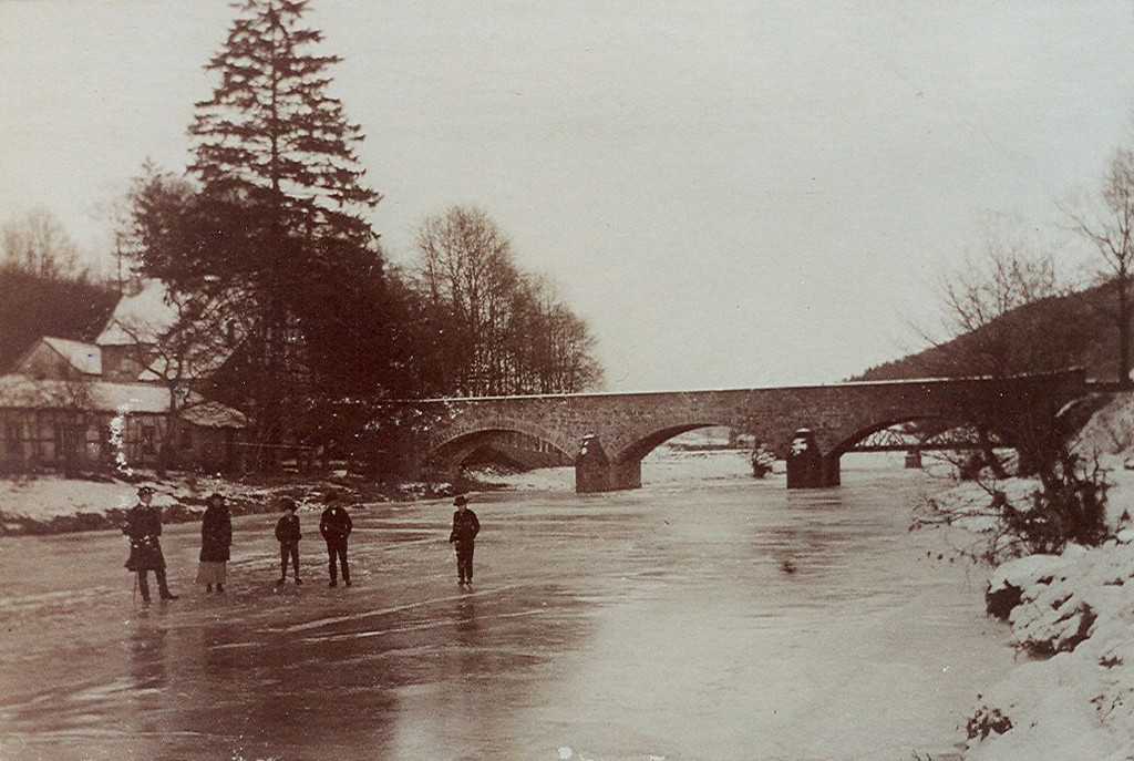 Historische Aufnahme (Ende 19./Anfang 20. Jahrhundert): Blick auf Schlittschuhfahrer auf der zugefrorenen Agger bei Engelskirchen-Loope. Im Hintergrund ist die alte Steinbrücke zu sehen.