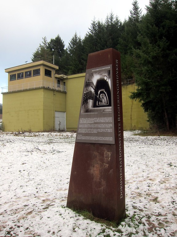 Steele mit Hinweistafel zur Dokumentationsstätte Regierungsbunker vor einem zu Wachzwecken ausgebauten Hochbunker des Regierungsbunkers Ahrtal an dessen westlichem Ausgang oberhalb von Marienthal (2015).
