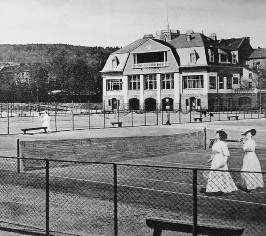 Damen beim Lawn-Tennis auf dem Sportplatz in der Bonner Reuterstraße (Aufnahme um 1910 in Blickrichtung Südwesten). Hinter dem Zaun der Turnplatz und im Hintergrund das 1906 erbaute Clubhaus des Bonner Eisklubs und spätere "Haus der Jugend".