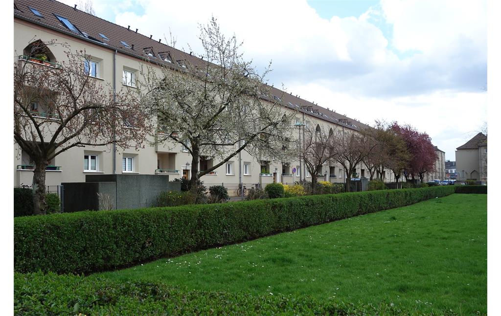 Köln-Mauenheim: Die dreigeschossige Blockrandbebauung aus Mehrfamilien-Reihenhäusern, umschließt große, begrünte Innenhöfe. Formschnitthecken und Baumreihen unterstreichen die formale, geometrische Gliederung (2016).