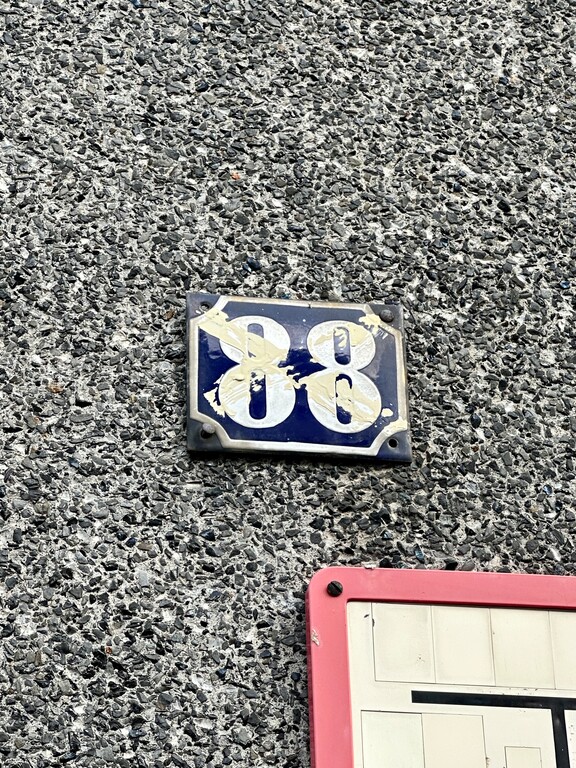 Die durchgestrichene Nummer 88 an der Hausfront erinnert an die vormalige Zählung der Hausnummern entlang der Hauptstraße. Diese begann bis in die 1960er Jahre erst an der Ortsgrenze zu Zündorf. Heute wird das Eckhaus als Bahnhofstraße Nr. 6a gezählt (2023).