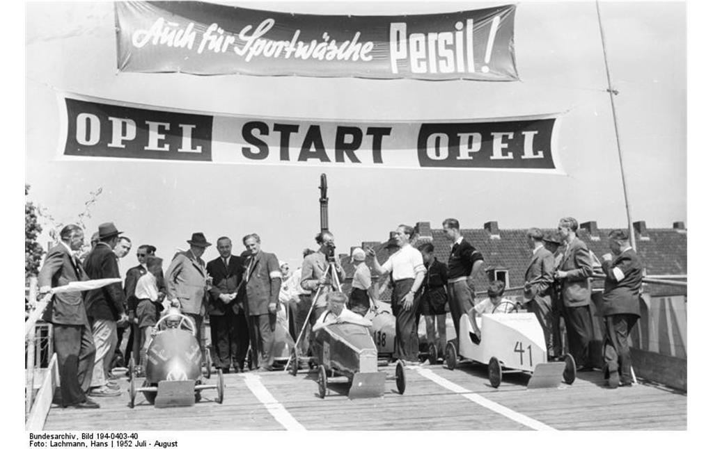 Start von drei Seifenkisten von einer für diesen Zweck erbauten Rampe zu einem Seifenkistenrennen in Duisburg (1952). Auf Fahnen wird für den Fahrzeughersteller "Opel" und das Waschmittel "Persil" geworben.