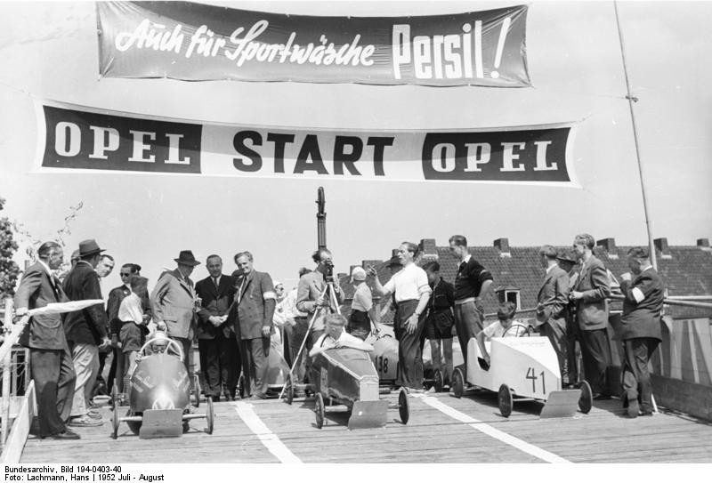 Start von drei Seifenkisten von einer für diesen Zweck erbauten Rampe zu einem Seifenkistenrennen in Duisburg (1952). Auf Fahnen wird für den Fahrzeughersteller "Opel" und das Waschmittel "Persil" geworben.