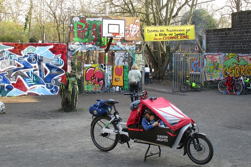 Der Eingang zum "Bauspielplatz Friedenspark" (Baui) im heutigen Kölner Friedenspark im Inneren Grüngürtel in Neustadt-Süd (2022). Auf einem Plakat wird der Spielplatz für Kinder und Jugendliche ausdrücklich als "Elternfreie Zone" deklariert.
