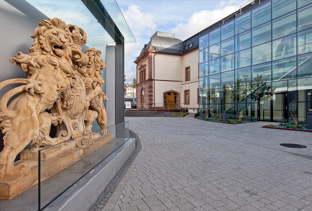 Links ist ein Sandsteinrelief von zwei Löwen mit dem königlich bayerischen Wappen in der Mitte zu sehen. Rechts die Glasfassade der modernen Erweiterung des Forums Alte Post in Pirmasens (2014).