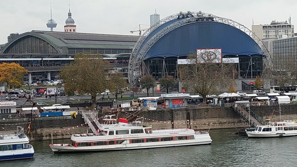 Das nach dem Kölner Volkssänger und Karnevalisten Wilhelm "Willi" Ostermann (1876-1936) benannte Personenfahrgastschiff auf dem Rhein, dahinter links der Kölner Hauptbahnhof und rechts der im Oktober 1996 eröffnete Musical Dome (2021).