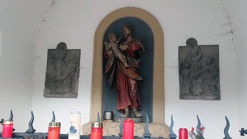 Figuren- und Bildschmuck in der Sankt-Josefs-Kapelle oberhalb von Bernkastel-Kues (2023): zentral eine Figur des heiligen Josef mit dem Jesuskind auf dem Arm, links und rechts davon Darstellungen der heiligen Familie.