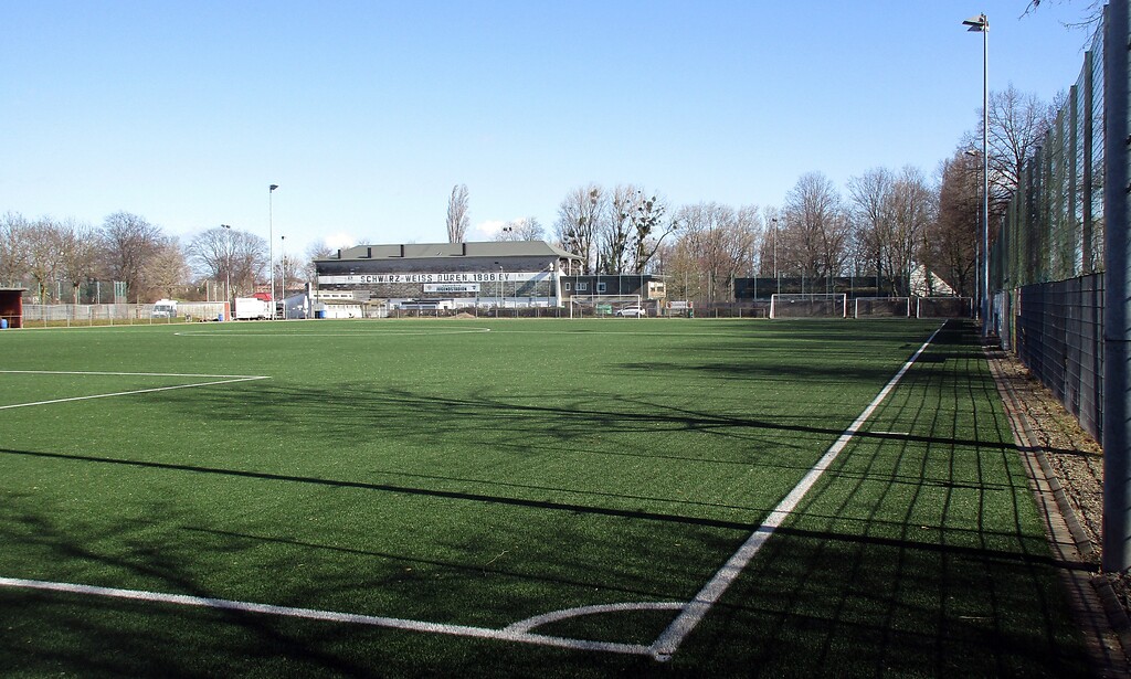 Blick über einen jüngeren Sportplatz in Düren-Rölsdorf auf die historische Sportanlage "Am Jugendstadion" im Hintergrund (2021).