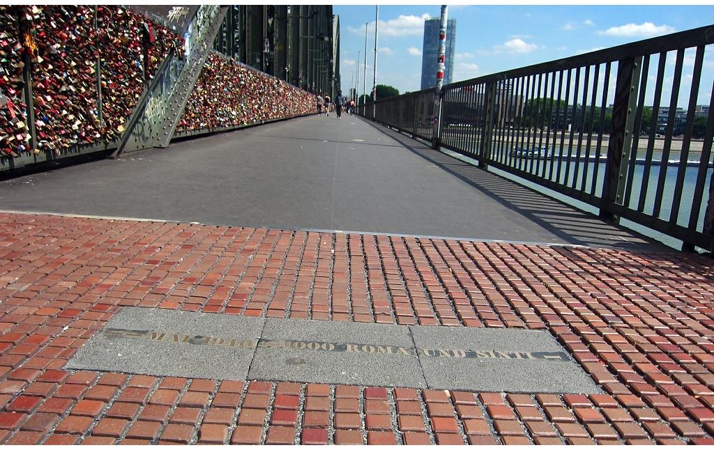 In das Pflaster eingelassene metallene Gedenkinschrift, die an die Deportation von Roma und Sinti über die Gleise von "Deutz-Tief" am Bahnhof Köln Messe/Deutz im Mai 1940 erinnert, am südwestlichen Zugang zur Hohenzollernbrücke (2015).