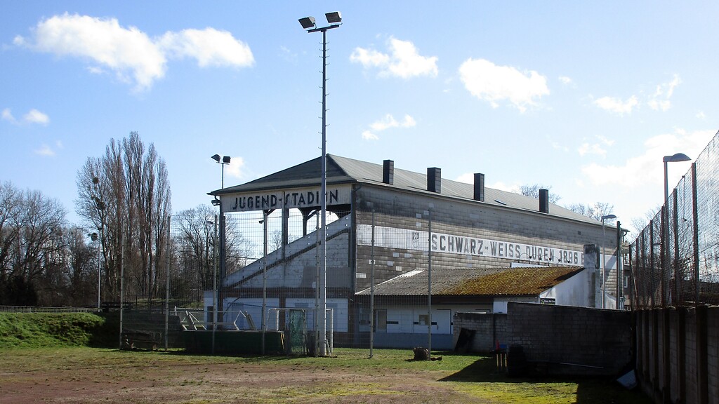 Blick auf die Rückseite der Tribüne an der 1921 eingeweihten Sportanlage "Am Jugendstadion" in Düren-Rölsdorf (2021), links im Bild ist der Wall der 1923 ergänzten Beton-Radrennbahn zu erkennen.