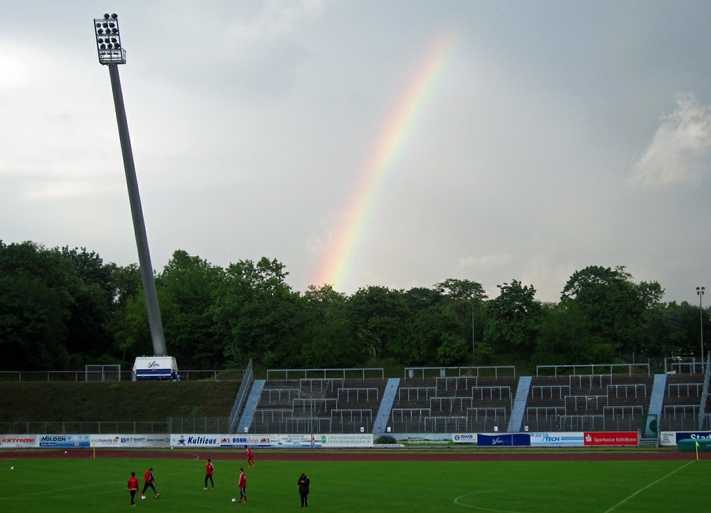 Die östliche Gegengerade des Sportstadions im Bonner Sportpark Nord von der Westtribüne aus gesehen, links im Bild ein Flutlichtmast und in der Mitte ein Regenbogen (2014)