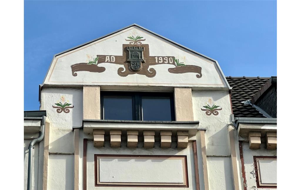 Das Haus Bahnhofstraße Nr. 6a in Porz-Mitte stammt aus dem Jahr 1907. Den Giebel zur Hauptstraße hin schmückt die Inschrift AD 1990, was auf eine Renovierung des Gebäudes im Jahr 1990 hinweisen dürfte, dazwischen ist das Porzer Wappen abgebildet (2023).