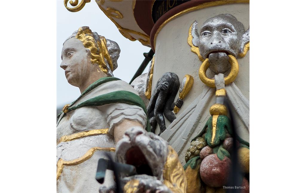 Detail am Petrusbrunnen auf dem Hauptmarkt in Trier (2022): Vier Statuen am Sockel stellen die Kardinaltugenden dar, hier links im Bild die Fortitudo (Tapferkeit und Stärke). Hinter ihrem Rücken am Brunnenstock betrachtet ein Affe sein Hinterteil im Spiegel.