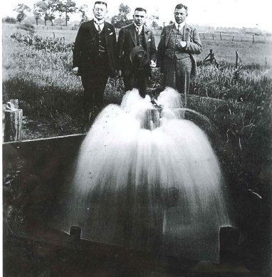 Drei Männer, die vor einer Wasserfontäne der Artesischen Quelle stehen. Die schwarz-weiß Aufnahme stammt aus der Zeit vor 1935.