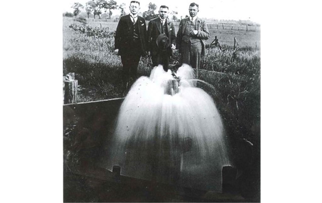 Drei Männer, die vor einer Wasserfontäne der Artesischen Quelle stehen. Die schwarz-weiß Aufnahme stammt aus der Zeit vor 1935.