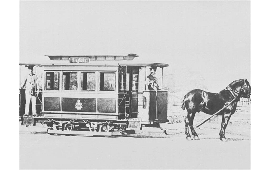 Historische Aufnahme eines Pferdebahn-Personenwagens mit stehendem Kutscher vorne an der Bremskurbel und Schaffner auf der hinteren Plattform (vor 1900).
