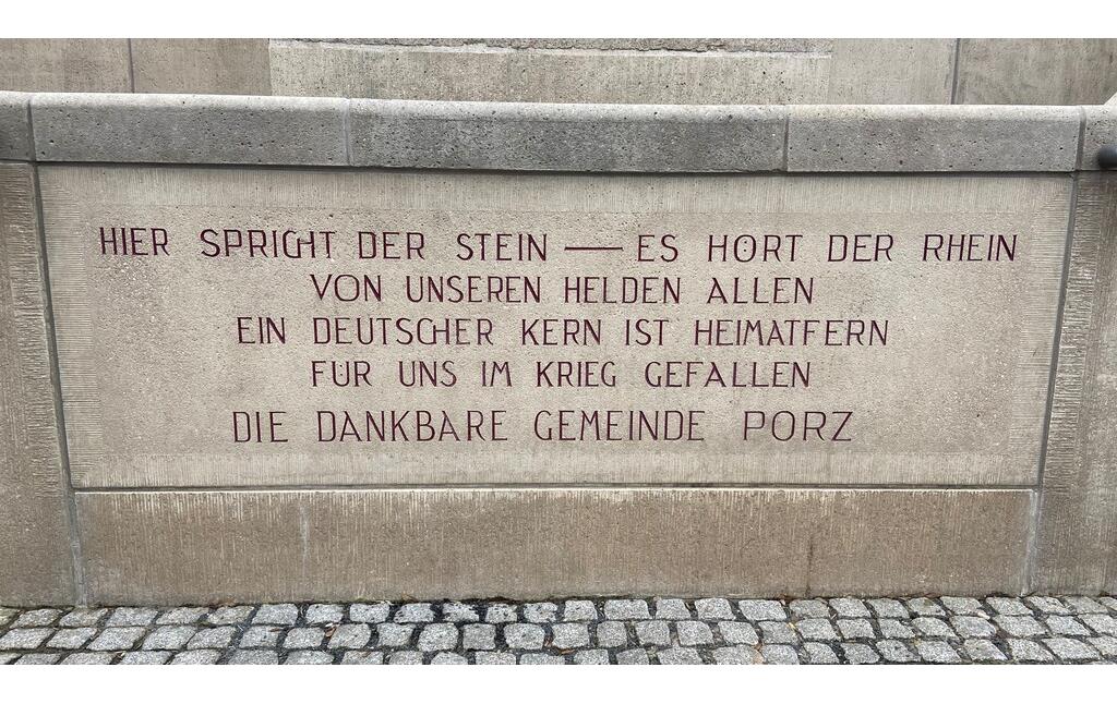 Die Inschrift der großen Treppenanlage am Porzer Rheinufer. In großen roten Lettern heißt es: "Hier spricht der Stein - es hört der Rhein. Von unseren Helden allen ein deutscher Kern ist heimatfern für uns im Krieg gefallen. Die dankbare Gemeinde Porz." (2023)