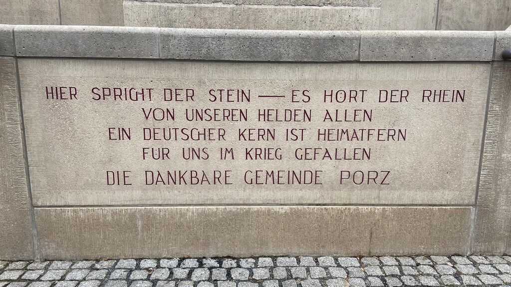 Die Inschrift der großen Treppenanlage am Porzer Rheinufer. In großen roten Lettern heißt es: "Hier spricht der Stein - es hört der Rhein. Von unseren Helden allen ein deutscher Kern ist heimatfern für uns im Krieg gefallen. Die dankbare Gemeinde Porz." (2023)