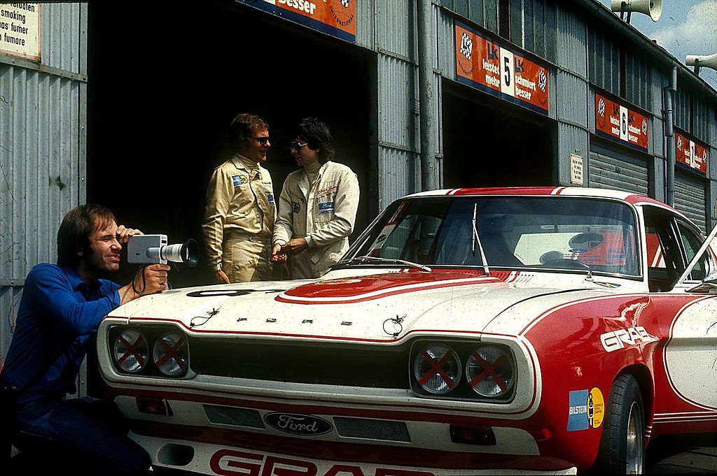 Ein Rennwagen des Typs Ford Capri RS mit den Autorennfahrern Karl-Ludwig Weiß (links) und Klaus Ludwig (rechts) sowie einem Kameramann im Fahrerlager des Nürburgrings (1973).