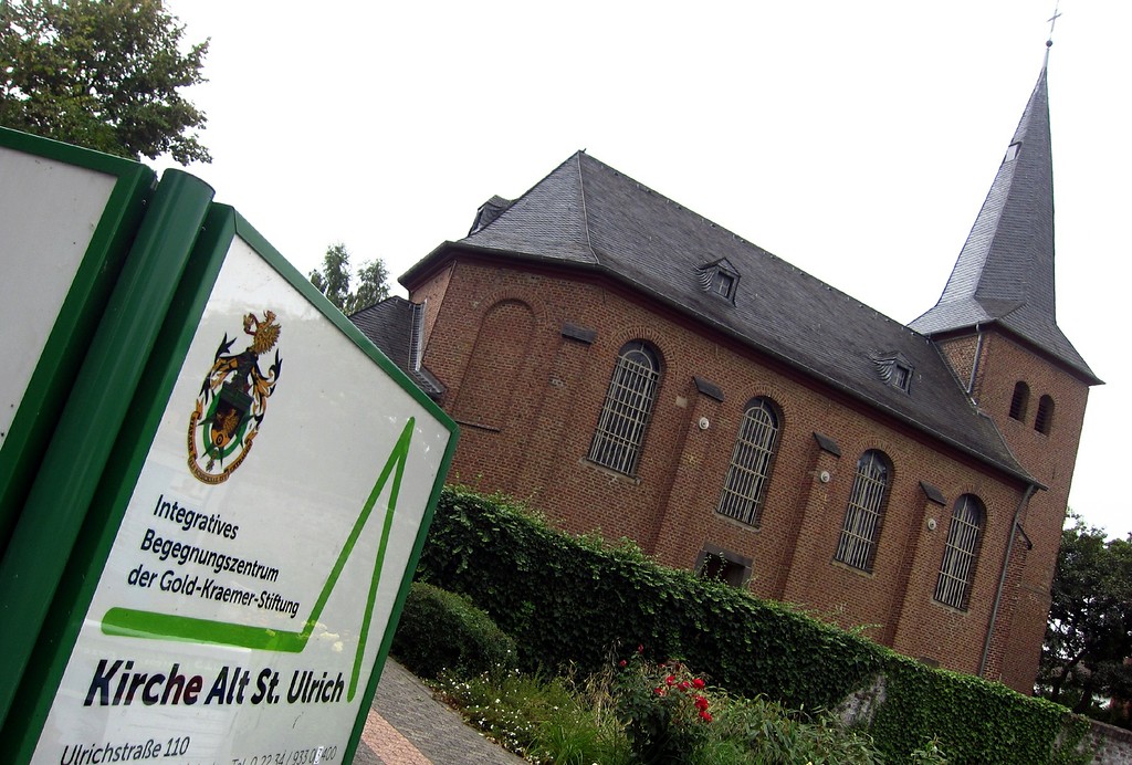 Die Kirche Alt St. Ulrich in Frechen-Buschbell von Südosten aus gesehen. Im Bild ist ein Hinweisschild auf das "Integrative Begegnungszentrum der Gold-Kraemer-Stiftung" zu sehen (2013)