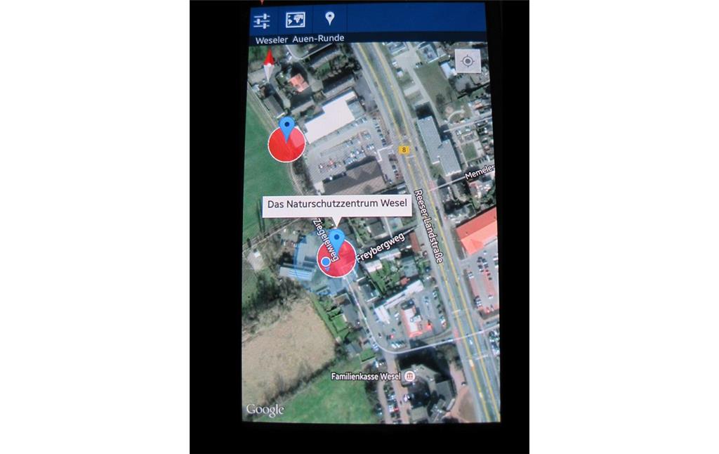 Bildschirm-Foto eines Mobilgerätes von der Anwendungssoftware "App in die Natur!". Es zeigt ein Luftbild von der Straße Freybergweg in Wesel mit dem Informationspunkt "Das Naturschutzzentrum". Der Informationspunkt ist Teil des Naturlehrpfad durch das Naturschutzgebiet Weseler Aue (2014).