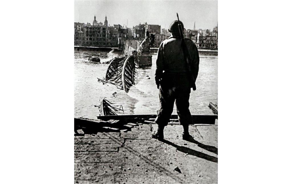 Aufnahme aus den letzten Tagen des Zweiten Weltkriegs in Bonn 1945: ein alliierter Soldat vor der am 8. März 1945 gesprengten Alten Rheinbrücke, im Hintergrund ist links die Bonner Namen-Jesu-Kirche zu erkennen.