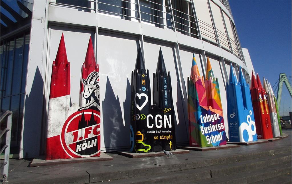Als Kölner Dom gestaltete Werbeobjekte am Schokoladenmuseum in Altstadt-Süd (2019), links mit dem Thema "1. FC Köln" und rechts davon mit dem Kürzel des Köln-Bonner Flughafens "CGN".